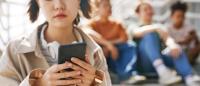 Closeup of teenage girl holding smartphone outdoors while sitting on metal stairs with group of teen friends in background